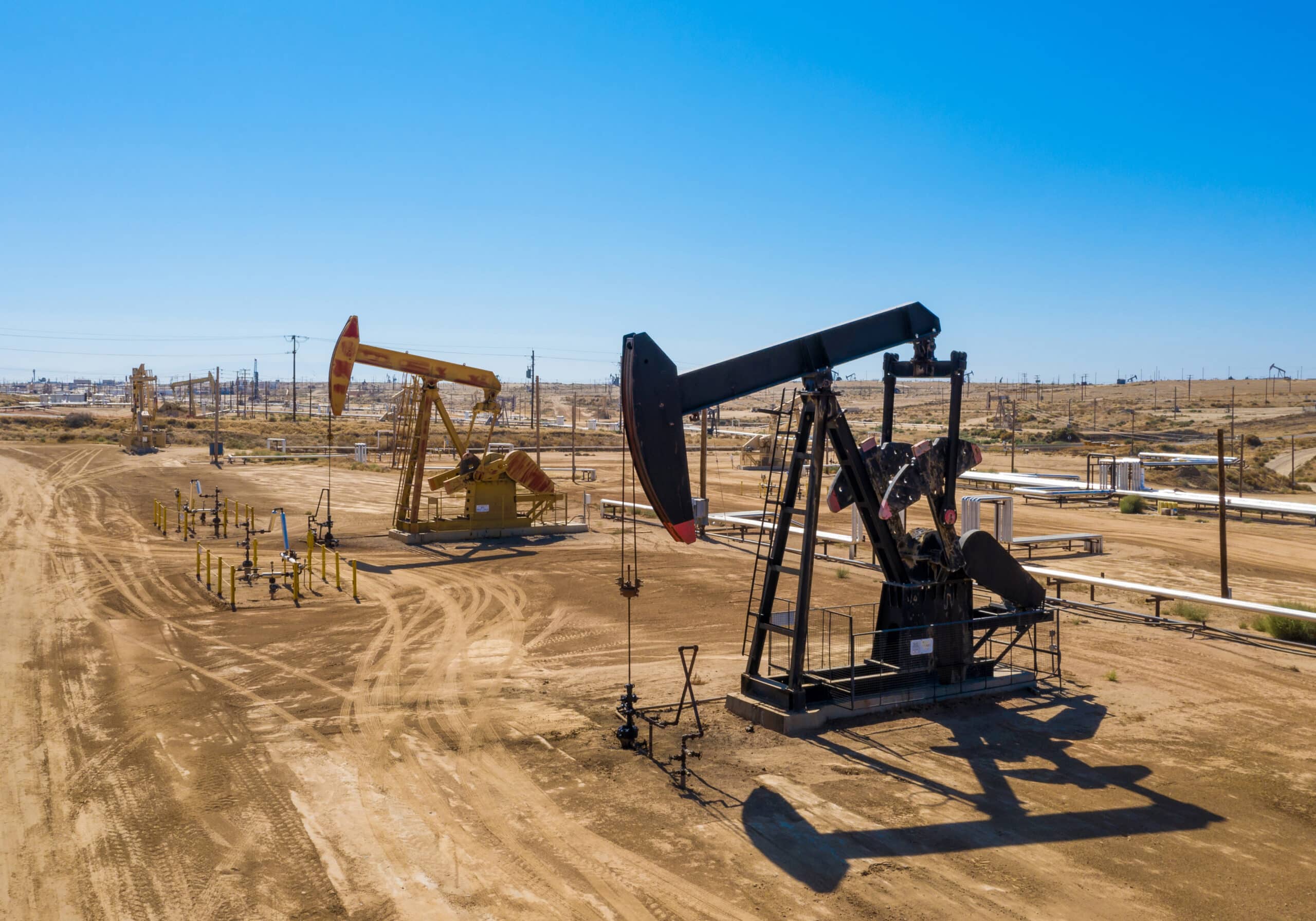 Two oil pumpjacks work in a dry, dusty oil field beneath a clear blue sky, with pipelines and industrial equipment behind them.