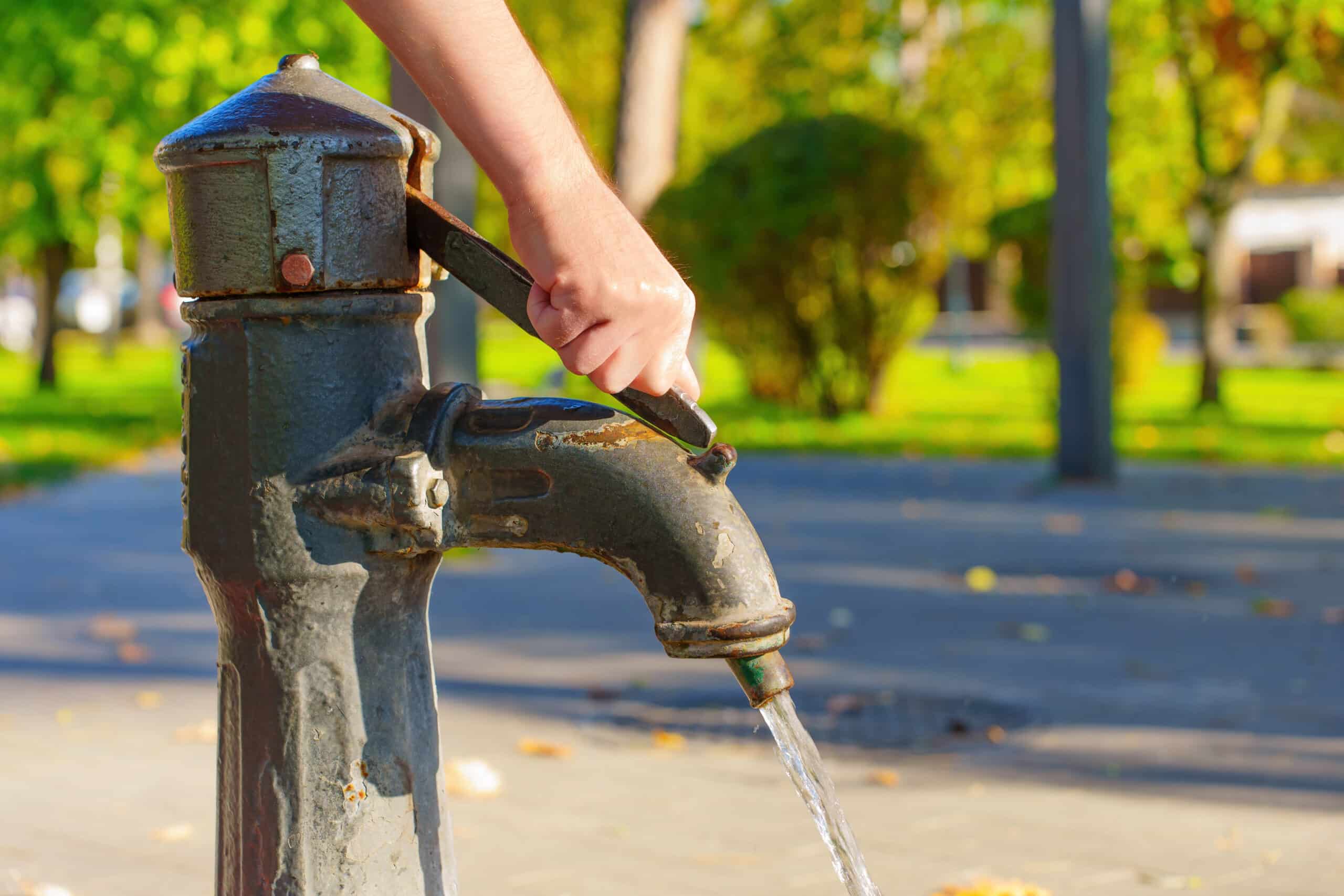 A hand works the handle of a vintage outdoor water pump as water pours out. Behind, a sunlit park features green trees and a path.