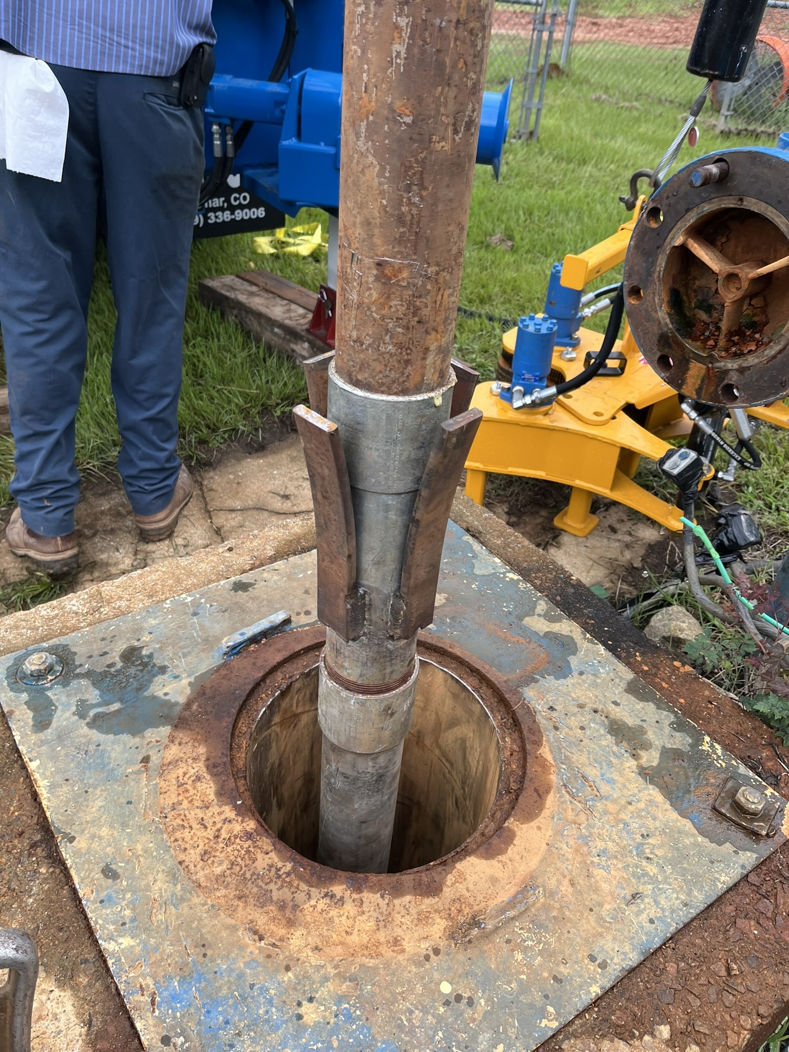 A metal pipe with a silver collar is being lowered into a round hole in a rusted plate outdoors as a worker and machinery stand nearby.