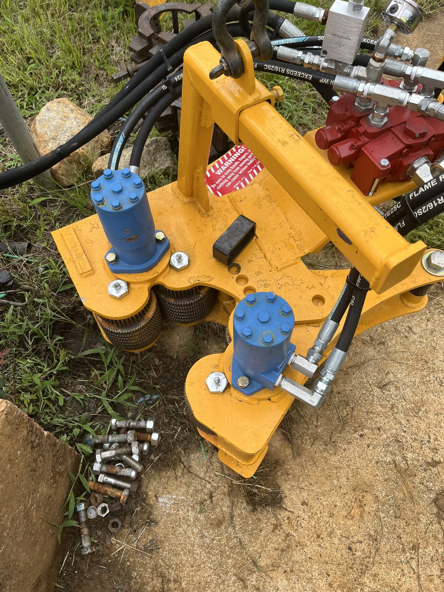 A yellow hydraulic tool with blue cylinders, black hoses, and metal parts lies on the ground by bolts, nuts, grass, and dirt.