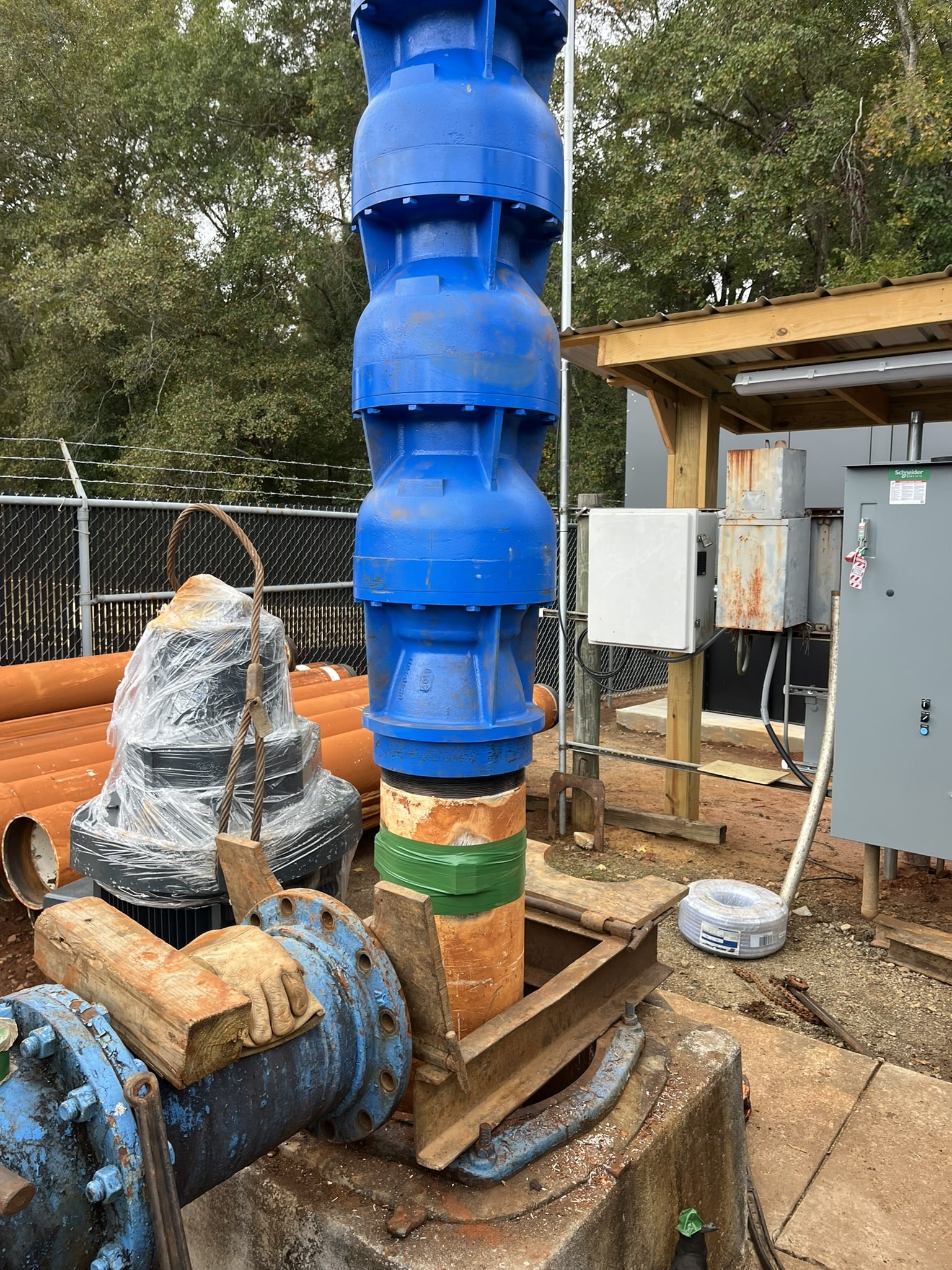 A large blue and orange industrial water pump sits on wooden blocks, surrounded by pipes, control panels, and trees outdoors.