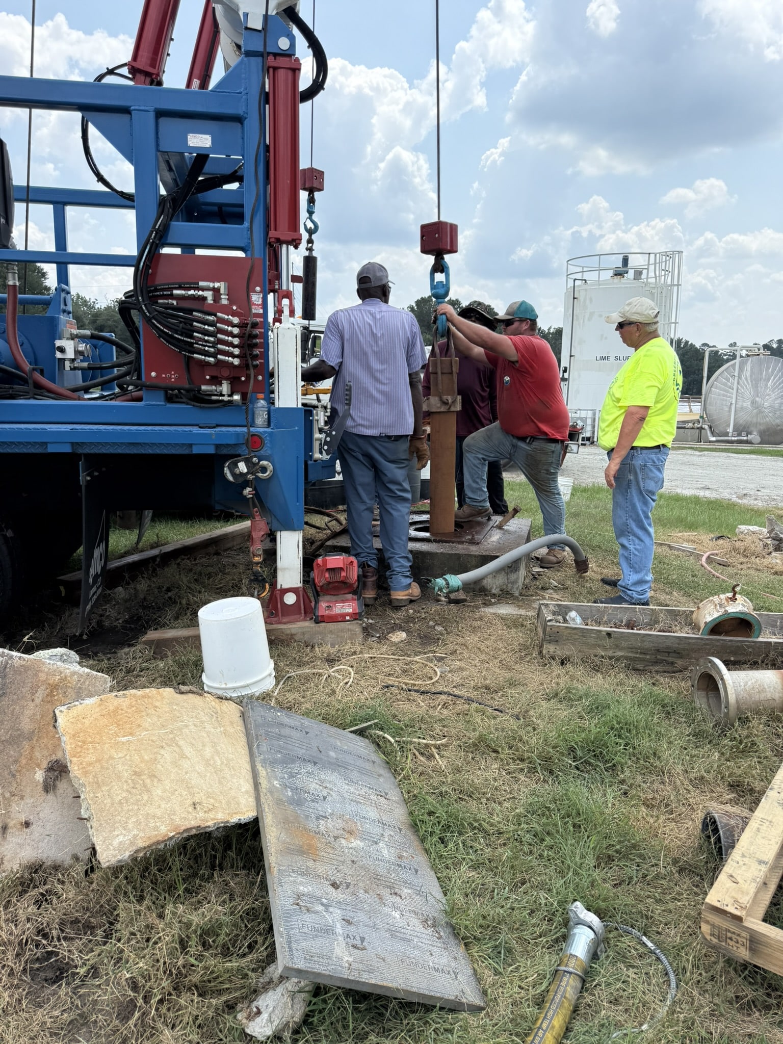 Four men at a drilling rig site handle equipment by a large machine, with tools and pipes on the ground under a partly cloudy sky.