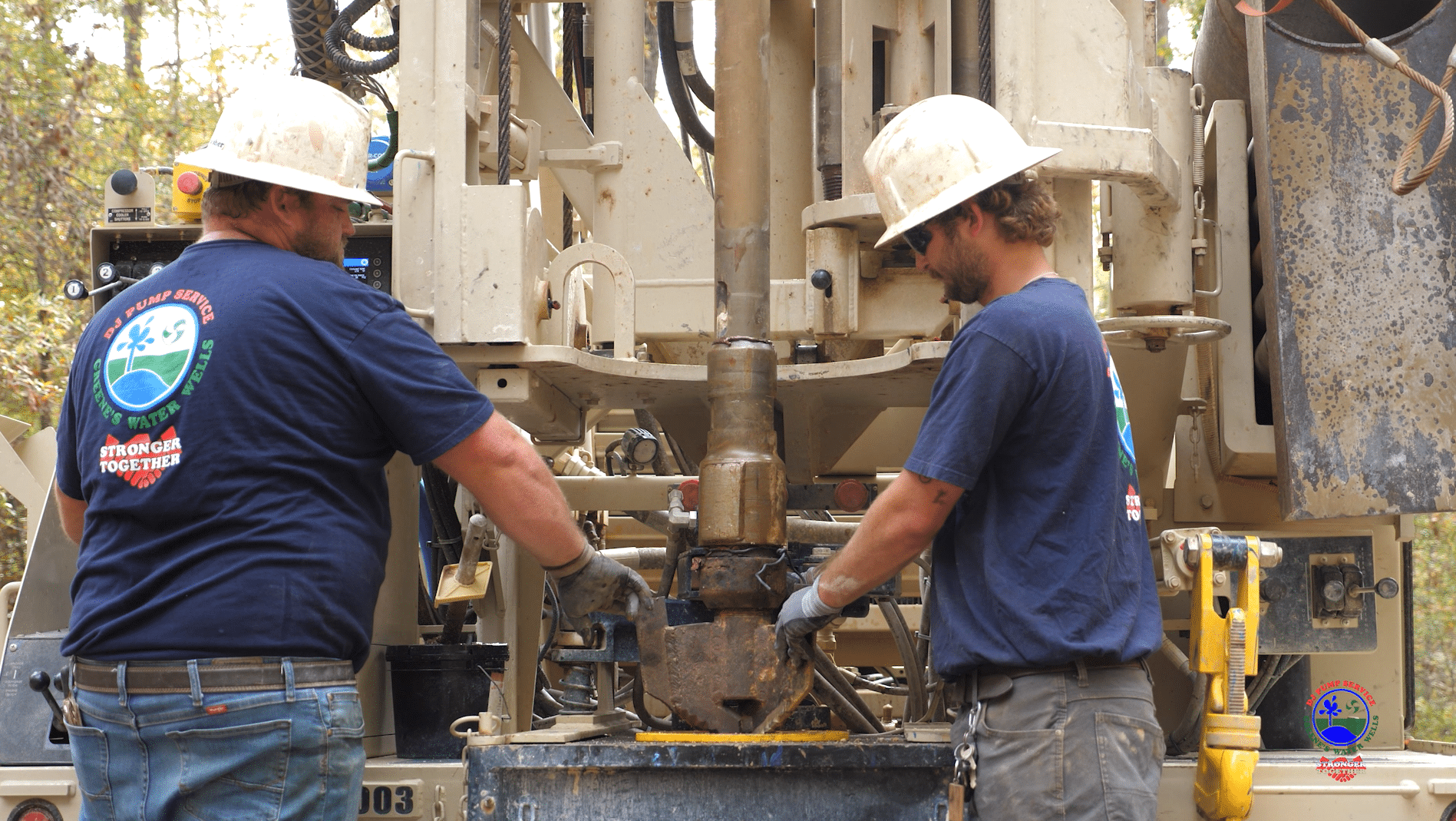Two workers in hard hats and gloves operate outdoor machinery, handling metal. Navy shirts show a circular logo. Trees, equipment behind.