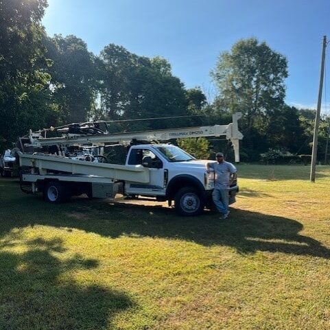 A utility truck with an extended boom arm is parked on a grassy field as two men stand nearby, surrounded by trees under a blue sky.