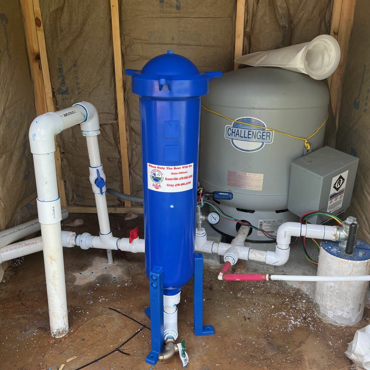 A blue water filtration unit and a large gray water tank labeled Challenger are installed in an unfinished wooden shed with exposed insulation and plumbing pipes.