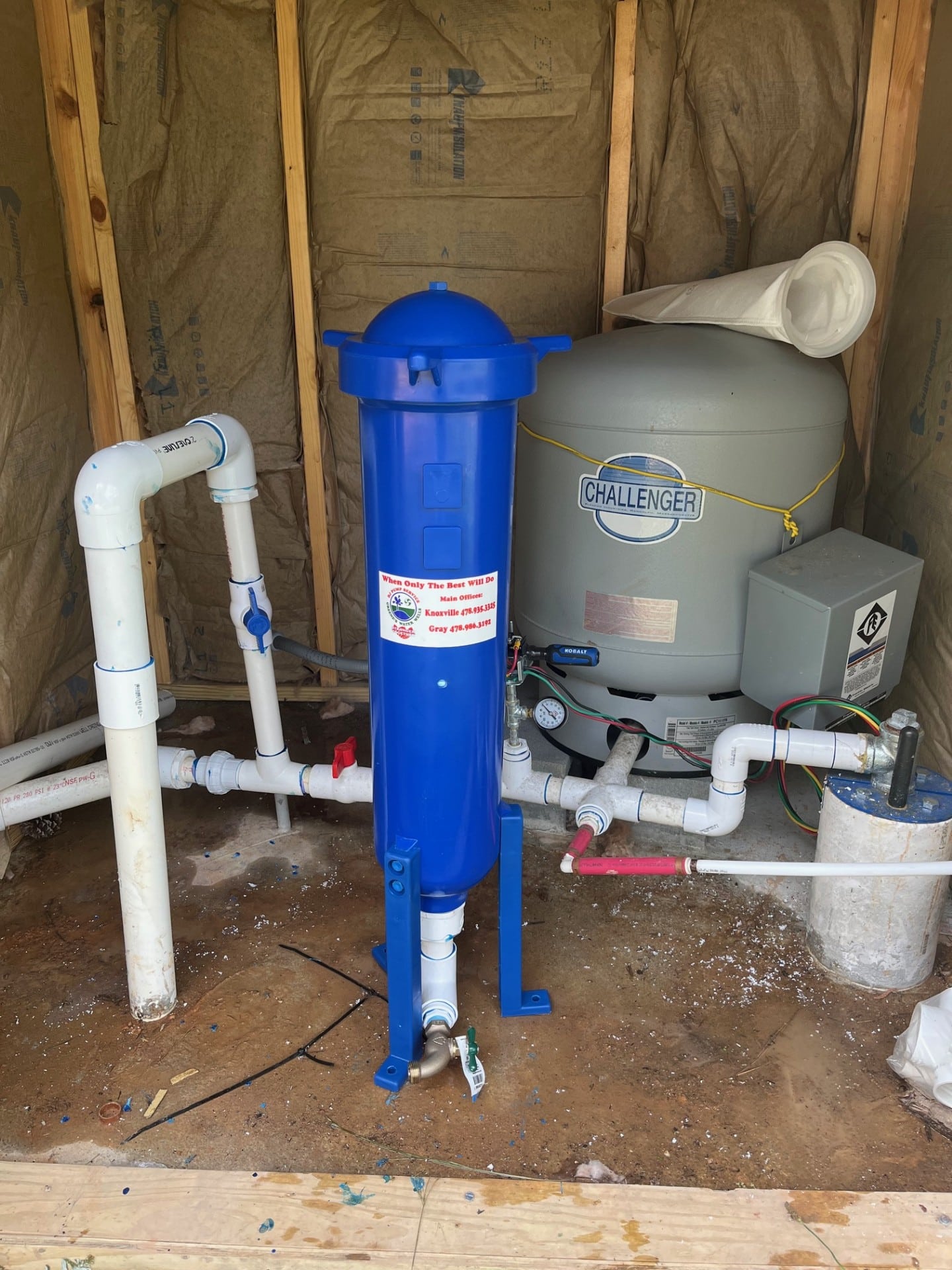 A blue water filtration system and a gray water pressure tank labeled Challenger inside a shed with exposed insulation and framing, with pipes and wires connected.