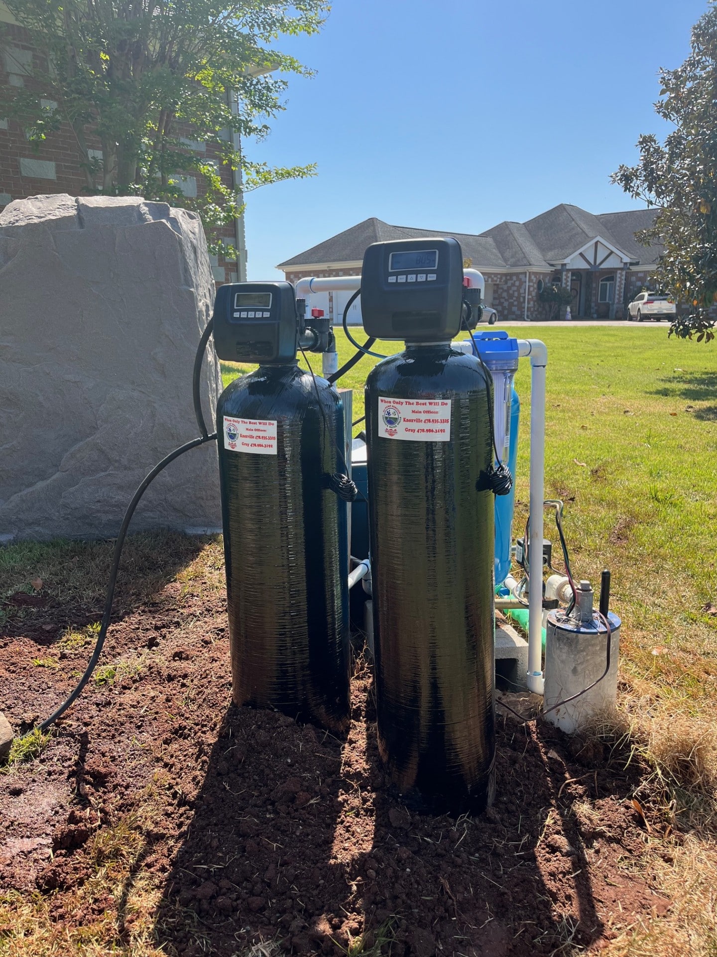 Two black water filtration tanks with digital panels outdoors on a lawn, pipes and wires connected, houses and trees in background.