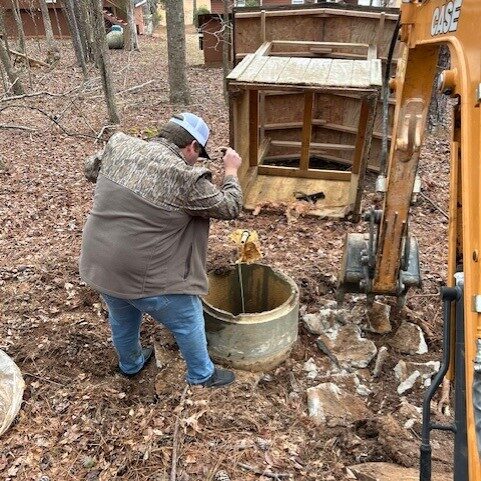 A man in a gray jacket and cap stands by a concrete well with tools amid trees, dirt, an excavator, and a wooden shed behind.
