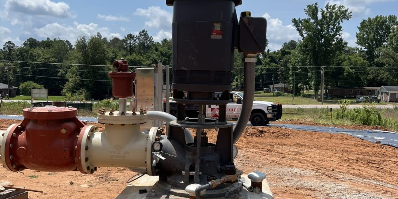 A large black industrial pump sits on a concrete slab at a construction site with red soil, and a white truck in the background.