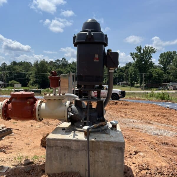 A large black industrial pump sits on a concrete slab at a construction site with red soil, and a white truck in the background.