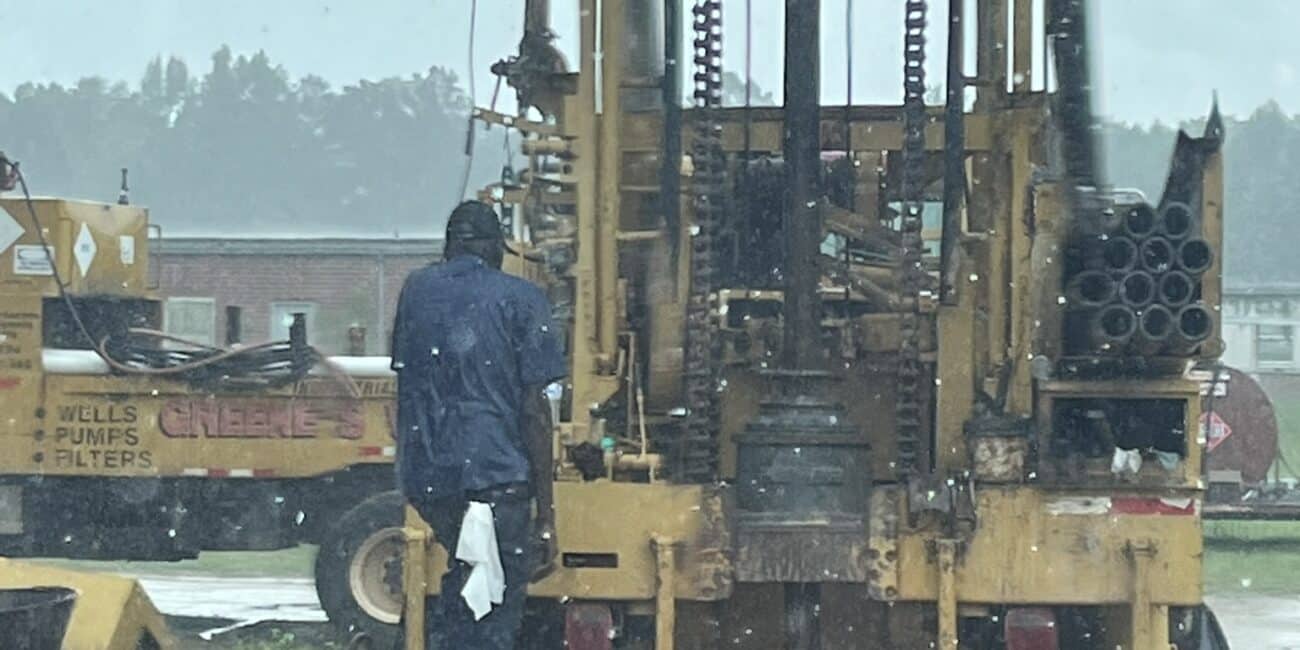 A person in a blue raincoat uses drilling equipment outdoors on a rainy day, with metal pipes, machinery, and wet ground nearby.