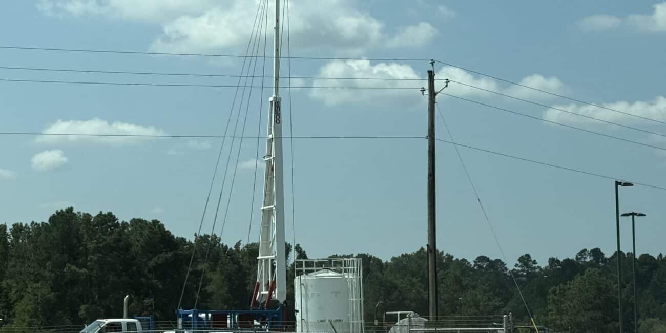 A construction site with a large white drilling rig, white tank, work truck, green pipes, and a Road Closed sign by a chain-link fence.
