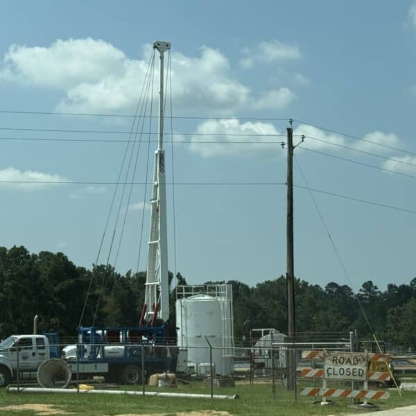 A construction site with a large white drilling rig, white tank, work truck, green pipes, and a Road Closed sign by a chain-link fence.