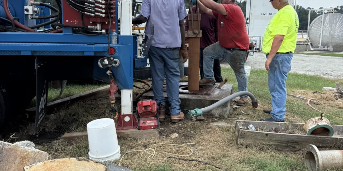 Four workers operate drilling gear at a grassy site with pipes, tools, and debris; one adjusts machinery as others assist under clouds.