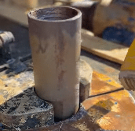 Close-up of a metal pipe clamped in a vise with visible paint marks and rust, blurred tools and equipment in the background.