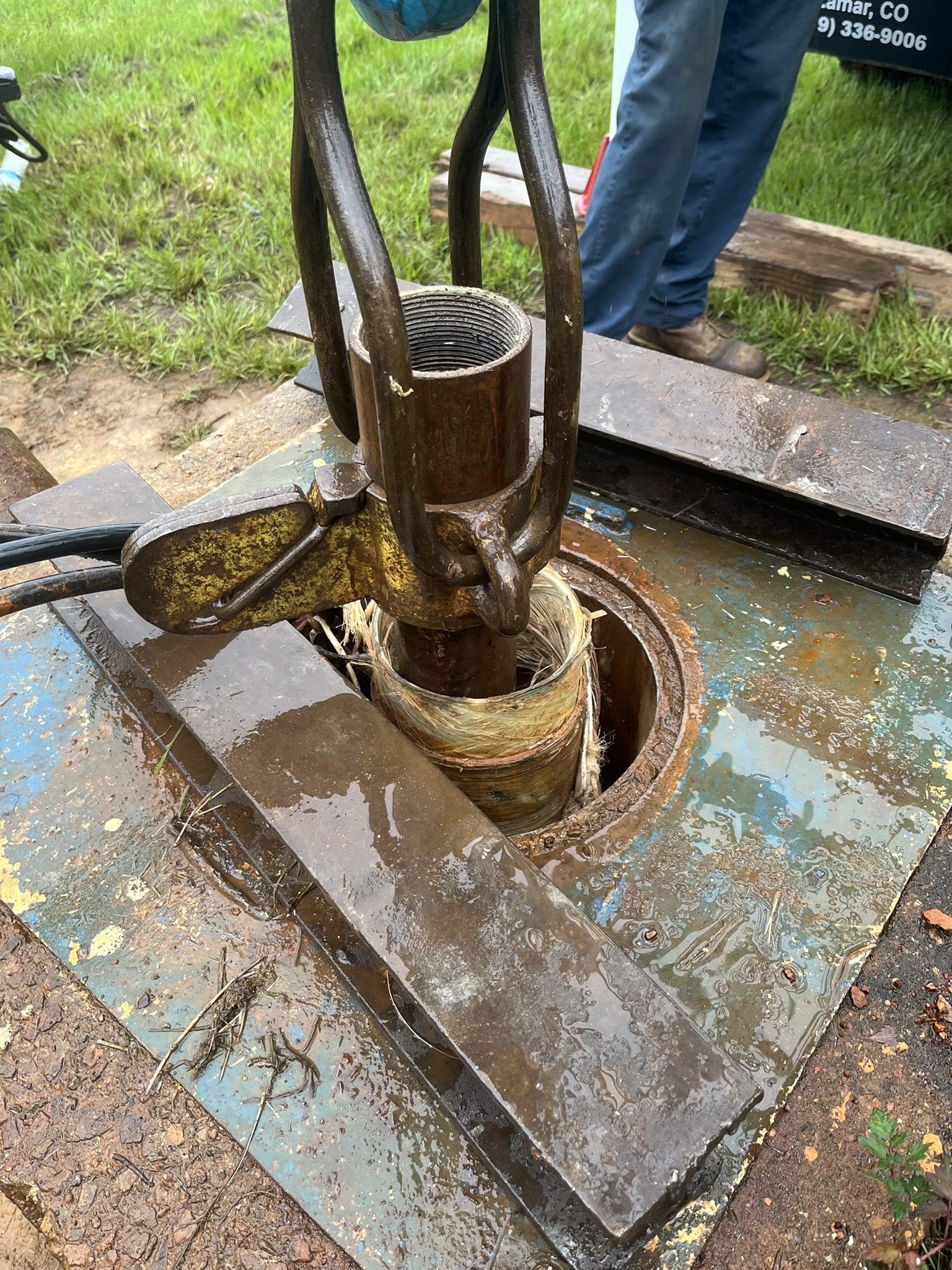 Close-up of well repair gear extracting a damaged pipe from a well, with a worker’s legs, grass, and repair tools visible in the background.