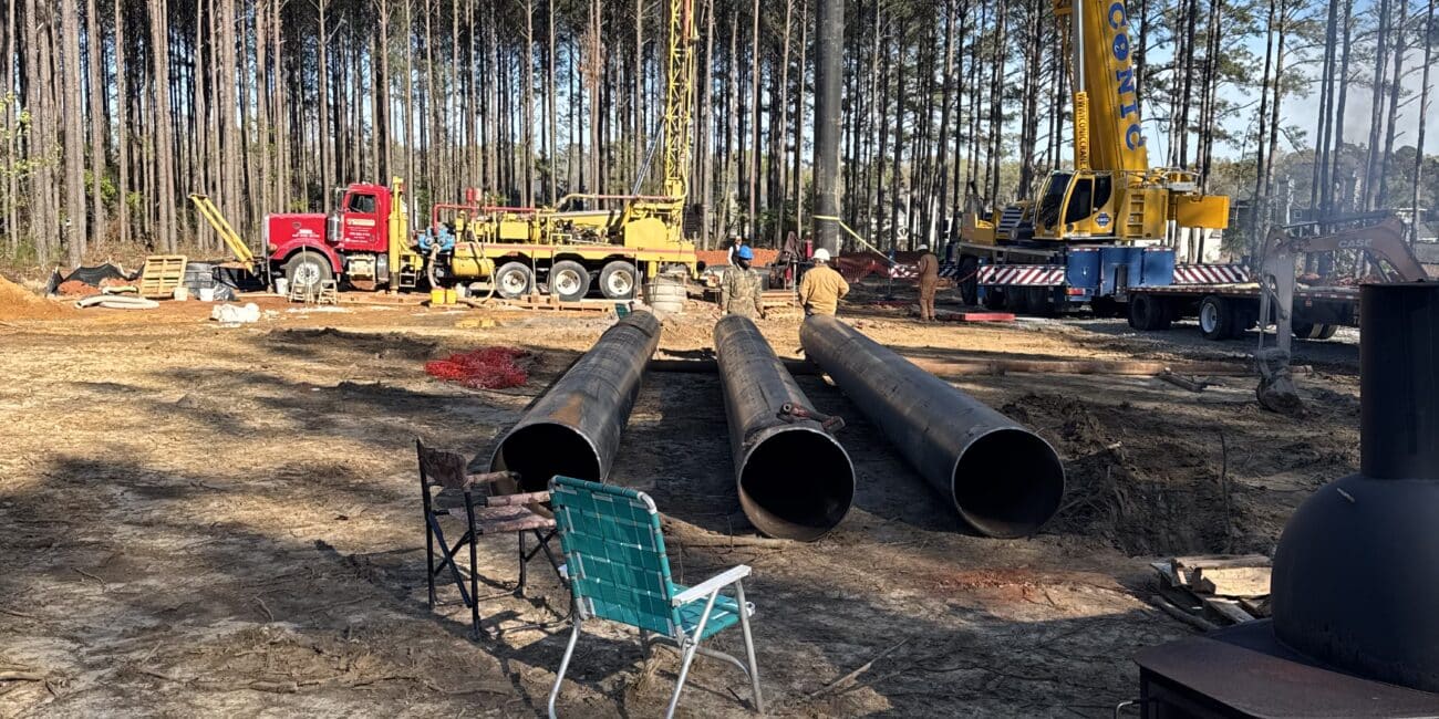 Construction site in woods with two cranes lifting pipes, a red truck, three metal pipes, folding chair, wood stove under blue sky.