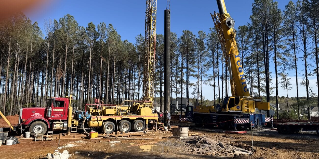Construction site with two large cranes, red and yellow trucks, workers, tall trees behind, and a finger over the top left corner.