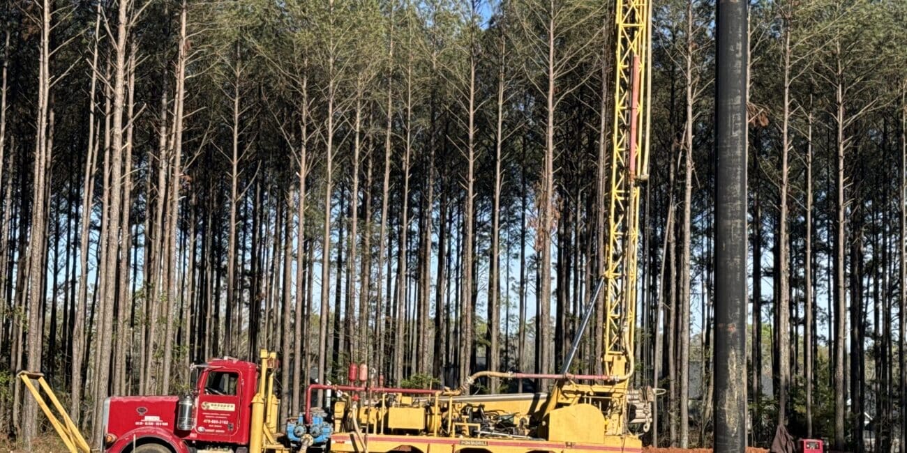 A large yellow drilling rig on a red truck sits in a pine forest clearing, with equipment and materials scattered around.